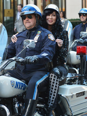 Cher chega na garupa da moto de um policial para se apresentar na TV